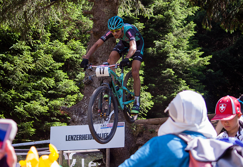 İsviçre'deki Lenzerheide UCI Dağ Bisikleti Dünya Kupası parkurunda 17 numaralı bisikletçi, çam ağaçları arasından geçerek havada zıplarken, bu zorlu dünyanın en ünlü bisiklet yarışları atmosferini yansıtıyor.
