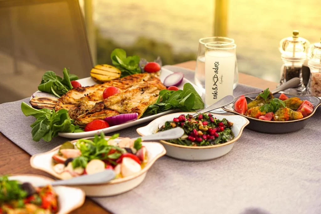 Güneşli bir akşamüstü manzarası eşliğinde, roka, domates ve kırmızı soğanla süslenmiş lezzetli ızgara balığın yanı sıra nar taneli salata, ahtapot salatası ve kabak çiçeği dolması gibi bol çeşitli mezelerin ve üzerinde Rakı & Adası yazan bir kadeh rakının servis edildiği zengin bir Türk deniz ürünleri sofrası.