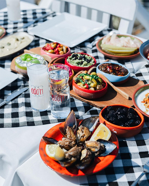 Siyah beyaz kareli bir masa, taptaze midye dolmalar, farklı renklerdeki küçük kaselerde zengin çeşitlilikte iştah açıcı mezeler ve üzerinde Ada'm yazan buzlu rakı kadehleriyle Bozcaada'nın gastronomik zenginliğini ve romantik kaçış rotası atmosferini yansıtan keyifli bir Ege sofrasını sergiliyor.