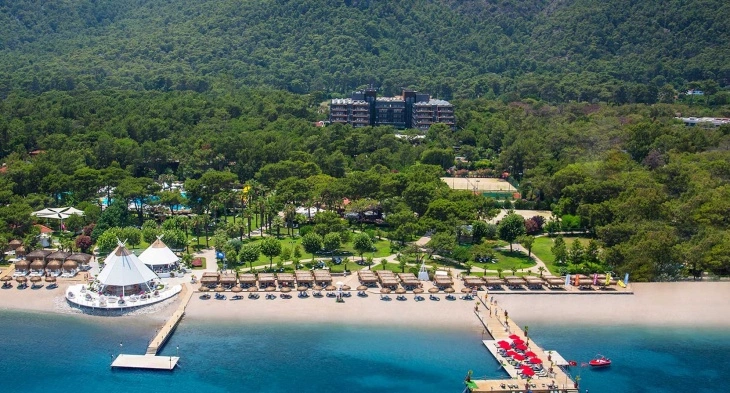 Yeşil ormanlarla çevrili, denize sıfır konumdaki bir aile otelinin geniş kum plajında sıralanmış şezlonglar ve şemsiyeler, beyaz konik çatılı mekanlar, masmavi denize uzanan iki iskele, yemyeşil bahçelerdeki havuzlar ve uzakta yükselen otel binasıyla Türkiye'nin sunduğu harika bir tatil manzarası.