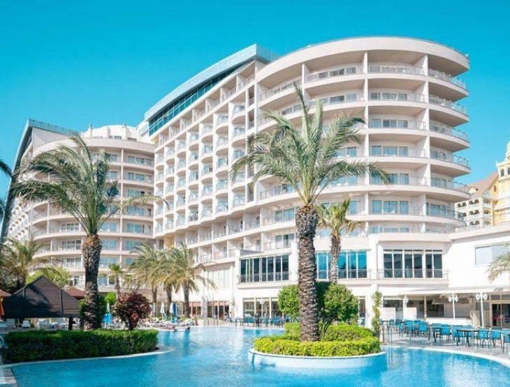 Büyük, modern ve kavisli mimariye sahip bir otel binası, sıcak Akdeniz iklimini yansıtan uzun palmiye ağaçları ve ferahlatıcı açık yüzme havuzu ile ailelerin keyifli bir tatil geçirebileceği Türkiye'nin en iyi aile otellerinden birinin sunduğu konforlu atmosferi gözler önüne seriyor.