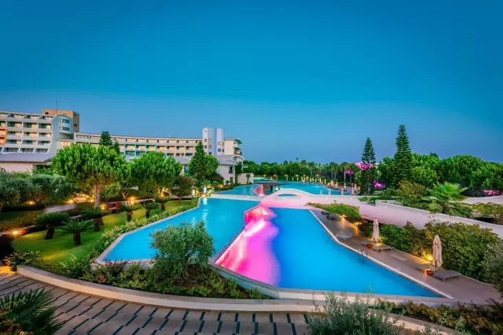 Akşamın loş ışıklarında, büyük bir aile otelinin önünde yer alan, pembe ve mor renkli ışıklarla aydınlatılmış bir su kaydırağı veya özel geçit içeren, köprülerle birbirine bağlı geniş, turkuaz renkli havuzlar ile yemyeşil bitki örtüsü ve ağaçlar, konforlu bir aile tatili vadeden görkemli bir manzarayı sergiliyor.