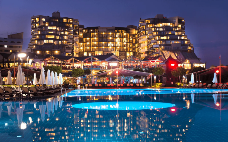 Akşamın karanlığında ışıl ışıl parlayan çok katlı bir otel kompleksinin önünde, mavi ışıklarla aydınlanmış geniş bir yüzme havuzu ve etrafındaki beyaz şemsiyelerle düzenlenmiş şezlonglar, Türkiye'nin en iyi aile otellerinde sunulan huzurlu bir akşam atmosferini yansıtıyor.