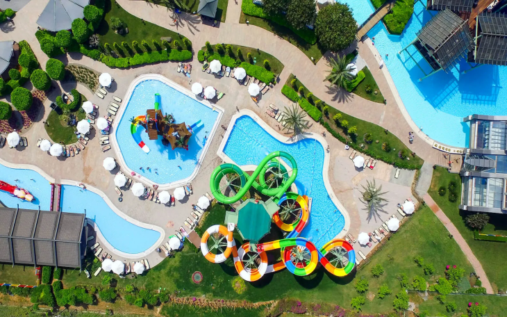 Türkiye'nin en iyi aile otellerinden birinin havadan görünümünde, ailelere yönelik eğlence olanaklarını vurgulayan rengarenk kaydıraklı büyük bir aquapark, içinde çocuk oyun alanı bulunan bir havuz ve güneşlenen misafirler için şezlonglarla çevrili berrak mavi havuzlar dikkat çekiyor.