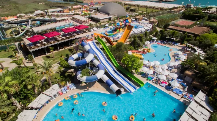 Türkiye’nin en iyi aile otellerinden birinde çekilmiş kuşbakışı bu canlı fotoğraf, farklı renklerdeki devasa su kaydıraklarıyla dolu büyük bir aquapark alanını, kalabalık havuzlarda yüzen çocukları ve yetişkinleri, çevresindeki şezlonglarda dinlenen tatilcileri ve yemyeşil palmiyelerle bezeli geniş bir dinlence alanını gözler önüne seriyor.