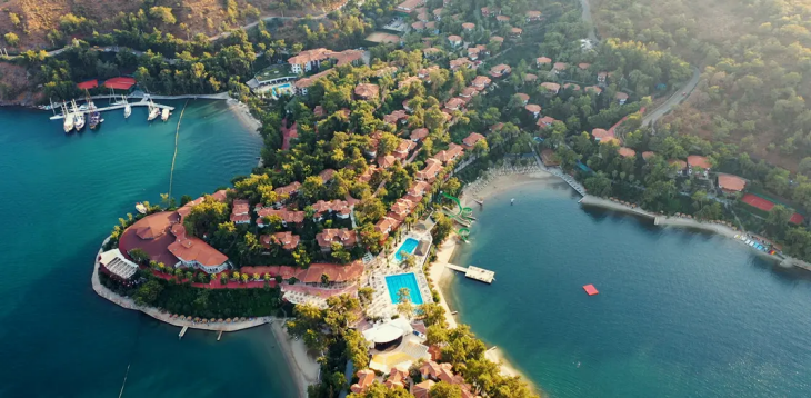 Yemyeşil ağaçlarla kaplı bir koyda, kırmızı çatılı binalar, birden fazla yüzme havuzu, kumlu plajlar, iskelede bağlı tekneler ve eğlenceli su kaydıraklarıyla dolu büyük bir tesisin kuşbakışı görüntüsü, Türkiye'nin en iyi aile otellerinden birinde unutulmaz bir tatil deneyimi sunuyor.