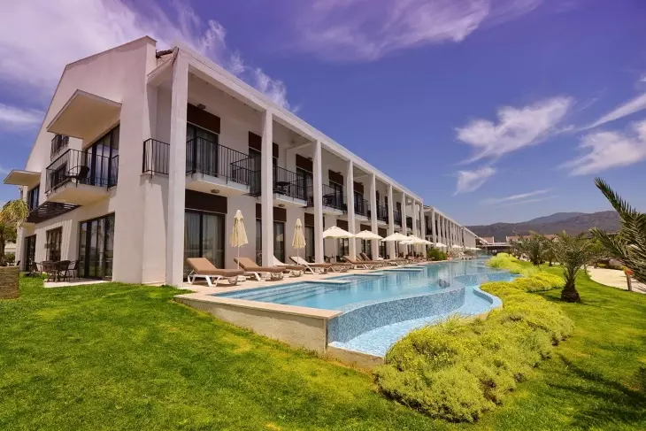 Beyaz modern binası, önündeki uzun, mavi mozaik kaplı havuzu, şezlongları, şemsiyeleri, yemyeşil çimleri ve arka plandaki palmiye ağaçları ile dağ manzarası sunan bu otel, Türkiye'nin en iyi aile otellerinden biri olarak konforlu bir tatil vaat ediyor.
