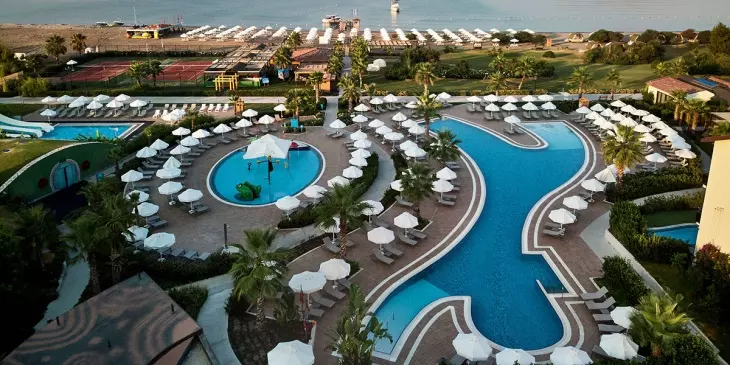 Türkiye'nin en iyi aile otellerinden birinde çekilmiş bu geniş açılı kuşbakışı görselde, kumlu plajı, çocuk aquaparkı ile süslenmiş havuzu, kıvrımlı ana havuzu ve çevresindeki yüzlerce şezlong ve şemsiye, ailelerin keyifli bir tatil geçirebileceği eğlence dolu olanakları gözler önüne seriyor.