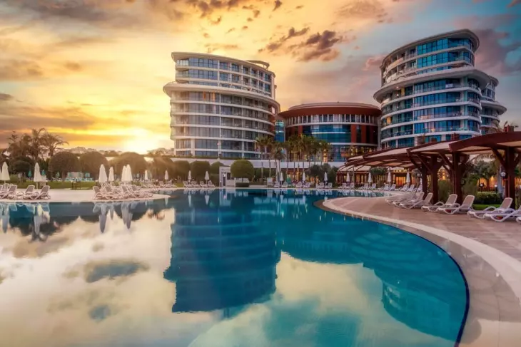 Gün batımının sıcak renkleri altında, modern mimariye sahip çok katlı otel binalarının ve yansımalarla parıldayan geniş bir yüzme havuzunun bulunduğu bu lüks tatil köyü, aile otelleri arasında unutulmaz bir konaklama deneyimi sunuyor.