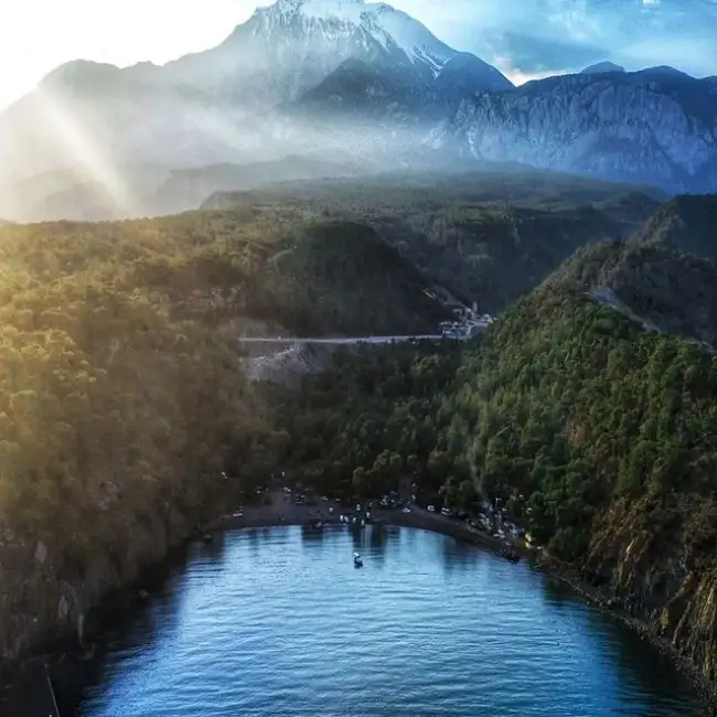 Antalya'nın gözde plajlarından Alacasu Cennet Koyu'nun havadan çekilmiş bu görüntüsü, yemyeşil tepelerin sardığı berrak turkuaz sularda demirlemiş bir tekneyle, kıyıda kamp kurmuş kalabalık grupları ve arka planda yükselen sisli, karlı dağları büyüleyici bir uyum içinde sunuyor.