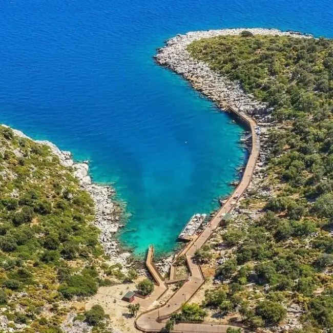 Antalya'nın en güzel plajları arasında yer alan Andrea Doria Koyu'nun kuşbakışı görünümünde, turkuaz renkteki berrak Akdeniz suyu, yemyeşil kayalık kıyılar, uzun ahşap bir iskele ve iskeleye bağlı birkaç tekne dikkat çekiyor.