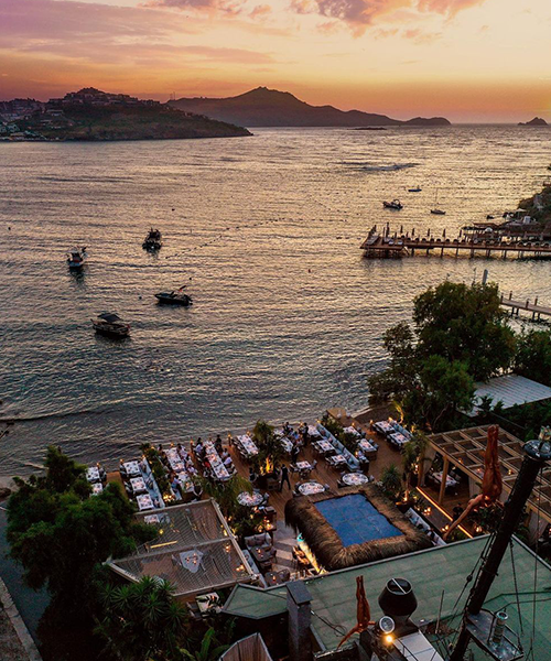 Yalıkavak'ta Ege Denizi'ne nazır konumlanan Azur Bodrum restoranı, gün batımının turuncu ve pembe tonlarıyla aydınlanan sahilde ve teknelerle dolu sakin denizde, şık masaları, iskelesi ve havuz başı lounge alanıyla Bodrum'un sofistike yeme-içme ve eğlence atmosferini gözler önüne seriyor.