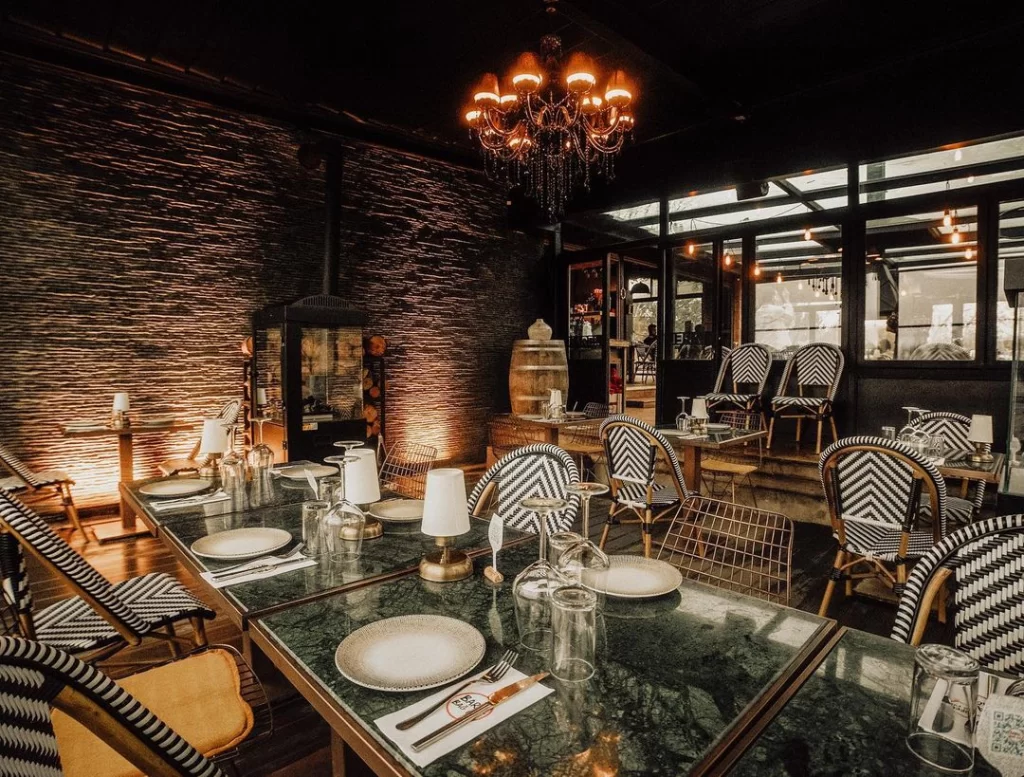 Loş ışıklı, şık bir restoran veya barın iç mekanında, koyu yeşil mermer üst yüzeyli masalar ve belirgin siyah beyaz zikzak desenli dokuma sandalyeler, arka plandaki dokulu koyu taş duvar, odunluklu modern bir şömine ve tavandan sarkan işlemeli zarif avize ile sıcak ve davetkar bir ambiyans hakim.