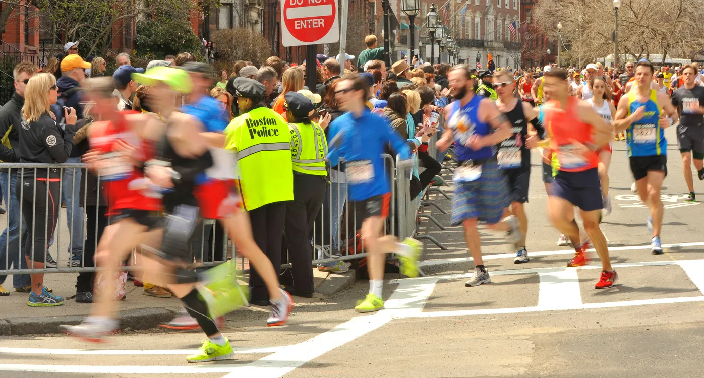 Tarihi Boston Maratonu'nda, kalabalık şehir caddelerinde ilerleyen, farklı yaş gruplarından binlerce koşucunun, güneşli bir günde, binalarla çevrili parkurda coşkuyla ve kararlılıkla kilometrelerce koştuğu dinamik bir sahne.
