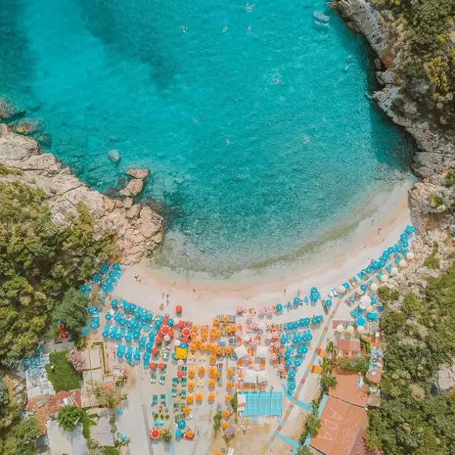 Hava fotoğrafında, yemyeşil kayalıklarla çevrili, kristal berraklığında turkuaz sulara sahip Büyük Çakıl Plajı, kıvrımlı kumlu sahilinde sıralanmış mavi, turuncu ve kırmızı renkli yüzlerce şemsiye ile şezlongların gölgesinde güneşlenen insanlarla dolu, burası Antalya'nın en güzel plajlarından biri olarak dikkat çekiyor.