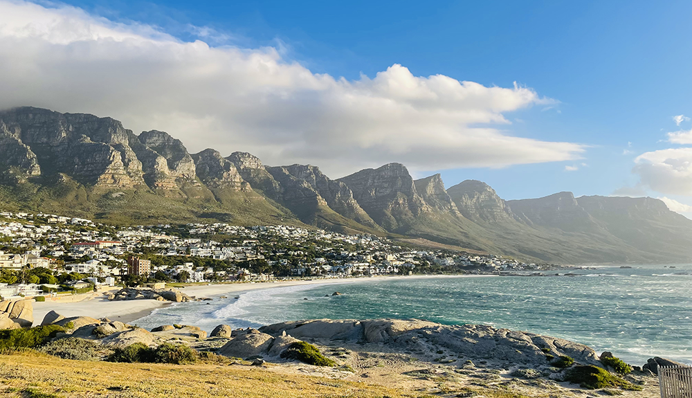 Cape Town'ın sahile yayılan yerleşim yerlerinin arkasında sarp kayalık zirveleri ve yemyeşil etekleriyle yükselen On İki Havari Dağları'nın, ön plandaki dalgalı turkuaz deniz ile geniş kumlu plajı ve bulutlu mavi gökyüzüyle bütünleşen muhteşem panoramik manzarası.