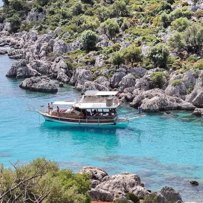 Antalya'nın berrak turkuaz sularında demirlemiş, yeşil bitki örtüsüyle kaplı kayalık kıyılarla çevrili bir koyda yolcularıyla keyifli vakit geçiren, çift katlı, ahşap bir gezi teknesi, bölgenin doğal güzelliklerini ve en güzel plajları deneyimleme fırsatını gözler önüne seriyor.