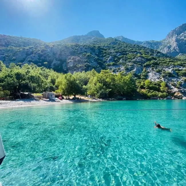 Parlak güneş altında, Antalya'nın en güzel koylarından Ceneviz Koyu'nun berrak turkuaz sularında bir kişinin yüzdüğü, arka planda yemyeşil ağaçlarla kaplı dik dağların ve küçük çakıllı plajın görüldüğü huzurlu bir manzara.
