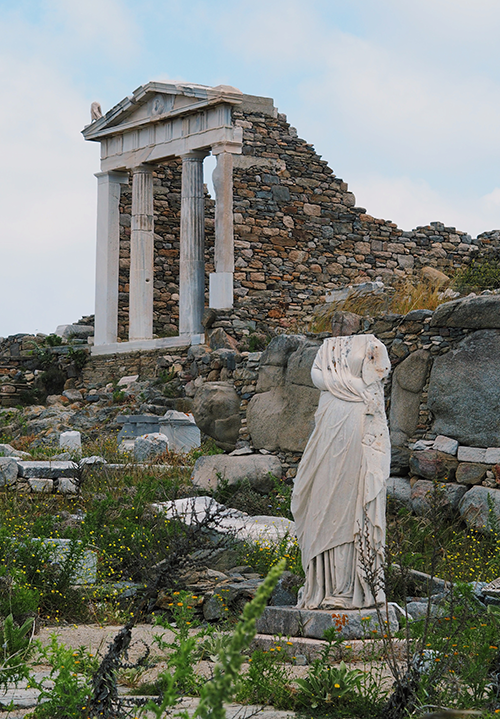 Mikonos seyahati sırasında ziyaret edilebilecek Delos Adası'ndan bir manzara; yemyeşil otlar ve moloz taşlarla bezeli arazide, başsız bir mermer heykelin önünde, arkada kısmen yıkılmış, beyaz sütunlu bir antik yapı ve taş duvarlar Ege'nin zengin tarihini gözler önüne seriyor.