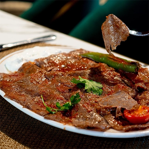 Kayseri'nin en beğenilen restoranlarından Elmacıoğlu İskender'in meşhur İskender kebabı tabağında, üzerinde bol domates sosu gezdirilmiş incecik et dilimleri, yanında tereyağlı yoğurt, közlenmiş yeşil biber ve taze maydanoz yapraklarıyla iştah açıcı bir sunum görülüyor, çatalda bir dilim et kaldırılıyor.