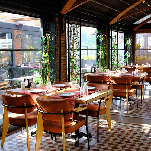 Kayseri'nin önde gelen restoranlarından birinin şık iç mekanını yansıtan bu geniş açılı görselde, tuğla duvarlar ve ahşap tavan detaylarıyla çevrili alanda, pencereden süzülen doğal ışık altında düzenlenmiş masalar, kahverengi deri kaplama sandalyeler ve geometrik desenli yer döşemeleri dikkat çekiyor.