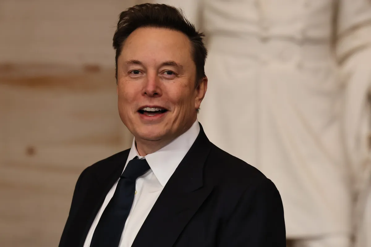 Koyu takım elbise ve lacivert kravat giymiş, hafif gülümseyen Elon Musk’ın iç mekânda çekilmiş portre fotoğrafı.