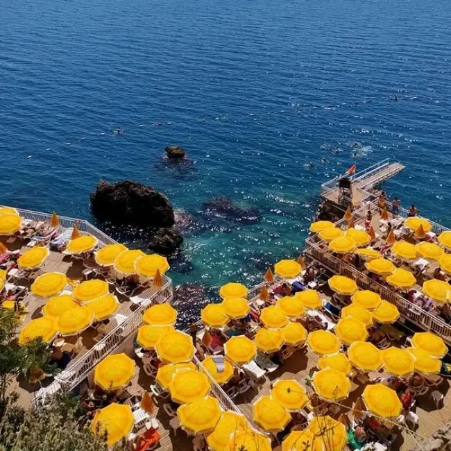 Antalya'nın falezler üzerine inşa edilmiş, berrak turkuaz sularına uzanan ahşap platformları, çok sayıda sarı şemsiye ve şezlongun altında güneşlenen ziyaretçileriyle öne çıkan İnciraltı Plajı'nın kuşbakışı, canlı bir manzarası.