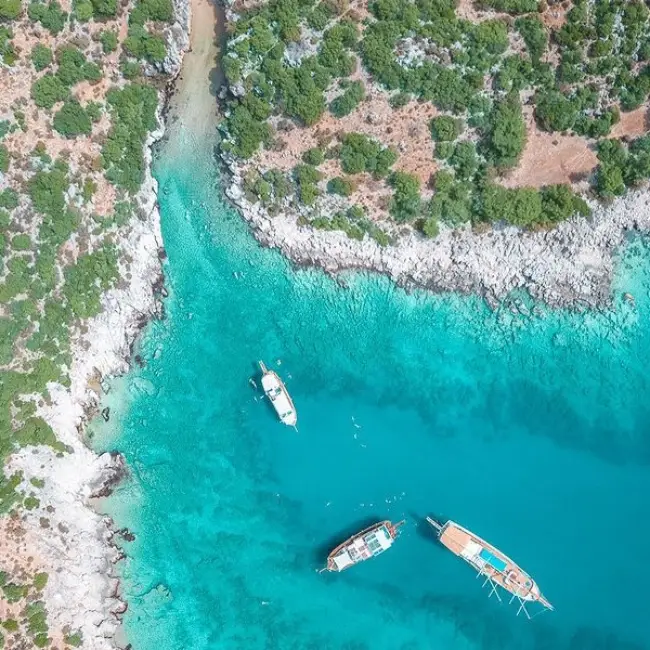 Antalya'nın en güzel plajlarından biri olan Kaş İnönü Koyu'nun kuşbakışı görünümünde, turkuaz renkli berrak sularında demirlemiş üç tekne, yemyeşil bitki örtüsüyle kaplı kayalık kıyı şeridi ve karaya doğru uzanan dar giriş dikkat çekiyor.