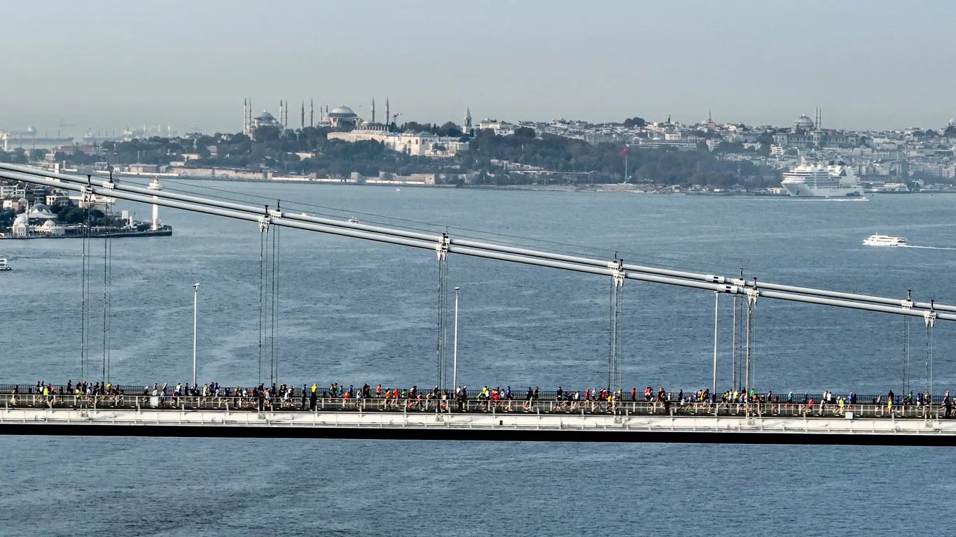 İstanbul Maratonu'nun heyecan dolu atmosferinde, farklı yaş gruplarından ve milletlerden yüzlerce koşucunun Boğaz köprülerinden birinde veya sahil şeridinde, bitiş çizgisine doğru kararlılıkla ilerlediği, coşkulu ve kalabalık bir an yansıyor.