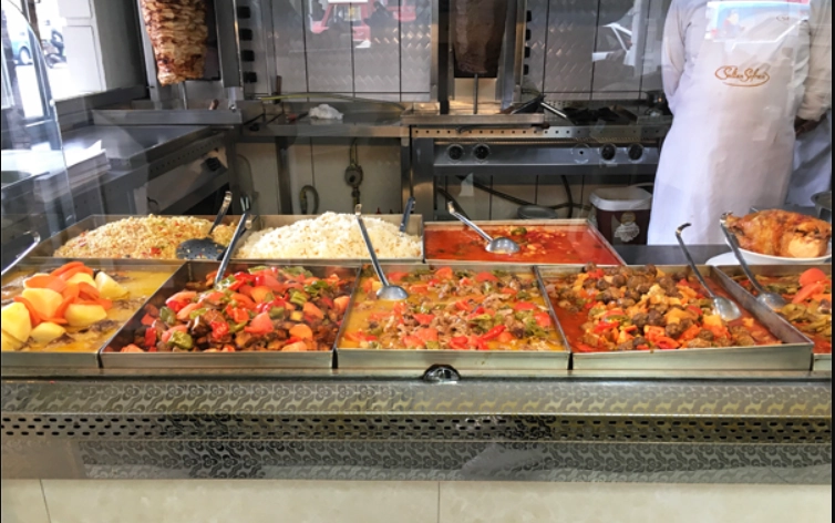 Kayseri'nin en beğenilen ev yemekleri restoranlarından Sultan Sofrası'nın açık büfe servis tezgahında, patatesli ve sebzeli etli yemekler, iki farklı pilav çeşidi, kırmızı soslu güveçler ve döner kebabın yanı sıra kızarmış bütün tavuğun da yer aldığı zengin menü sergileniyor; arka planda ise beyaz önlüklü bir aşçı görülüyor.