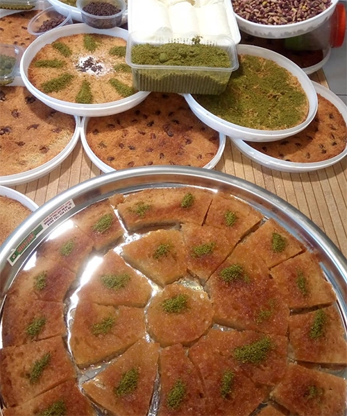 Kayseri'nin beğeni toplayan restoranlarından Kebapçı Şakir'in öne çıkan cevizli tahinli kadayıfının dilimlenmiş haliyle metal bir tepside sergilendiği, arka planda Antep fıstıklı kadayıfların, tahinli tatlıların ve rulo sütlü tatlıların beyaz plastik kaplarda hazırlandığı zengin bir tatlı sunumu.