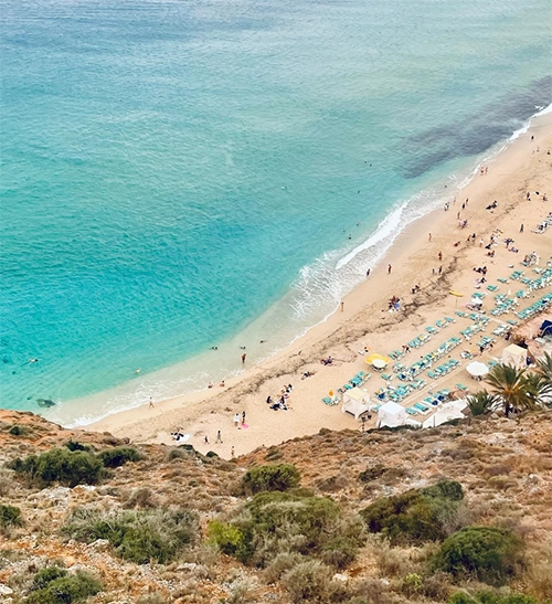 Antalya'nın en güzel plajlarından Kleopatra Plajı'nın kuşbakışı görüntüsünde, berrak turkuaz sulara sahip geniş kumsalda güneşlenen ve denize giren çok sayıda tatilci, düzenli sıralar halinde dizilmiş şezlong ve şemsiyeler, sağ alt köşede palmiye ağaçları ve ön plandaki kayalık, çalılık yamaç öne çıkıyor.