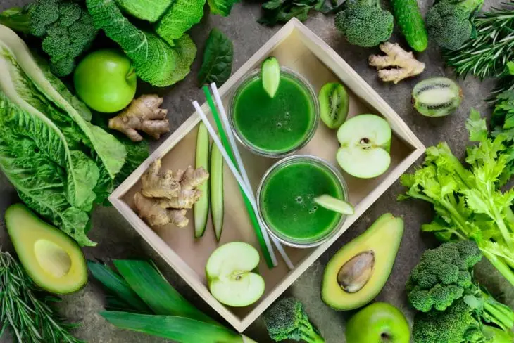 Ispanak, avokado, yeşil elma ve zencefil gibi malzemelerle hazırlanmış iki bardak yeşil smoothie, doğal malzemelerle çevrili şekilde servis ediliyor.