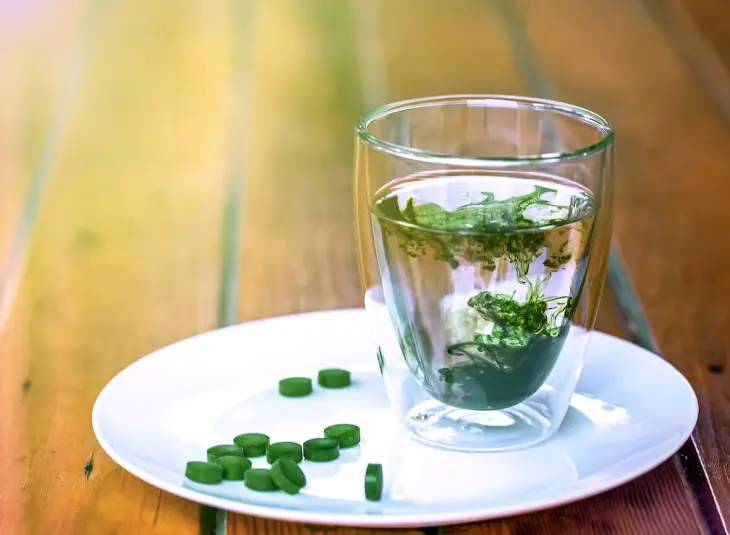 Beyaz tabakta yer alan spirulina tabletleri, cam bardakta su içinde çözünerek koyu yeşil rengini suya bırakıyor.