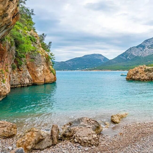Antalya'nın en güzel plajlarından biri olan Korsan Koyu'nun göz alıcı manzarasında, yemyeşil bitki örtüsüyle kaplı yüksek kayalıklar arasında uzanan cam gibi berrak turkuaz sular, küçük bir teknenin yüzdüğü huzurlu bir koy ve uzakta yükselen heybetli dağlar, çakıllı kıyıyı tamamlıyor.