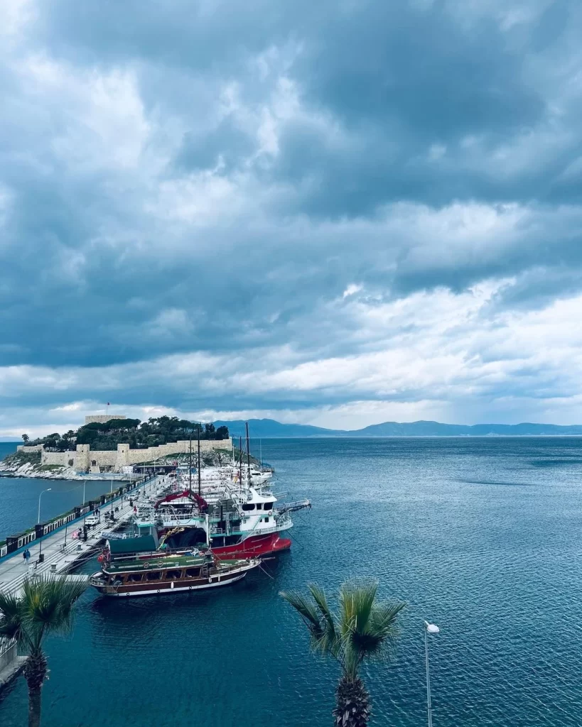 Kuşadası limanında, iskeleye bağlı farklı boyutlarda gezi tekneleriyle dolu, arka planda yemyeşil ağaçlarla süslenmiş tarihi Güvercinada Kalesi'nin yükseldiği, üzerinde dramatik koyu bulutların süzüldüğü geniş bir deniz ve gökyüzü manzarası.