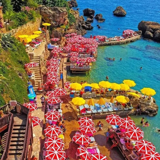 Antalya'nın eşsiz güzellikteki plajlarından Mermerli Plajı'nın falezlerin altında yer alan, berrak masmavi sularla buluşan kayalık kıyısında, kırmızı-beyaz ve sarı şemsiyelerle dolu şezlonglarda dinlenen ve denizin tadını çıkaran kalabalık bir grup insan kuşbakışı görünümde.
