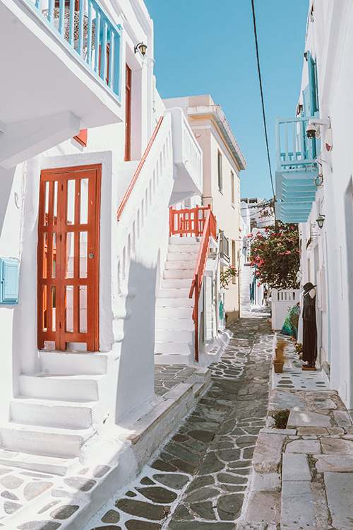 Mikonos'un kalbi sayılan Mykonos Town'ın dar, taş döşeli sokaklarından birinde, canlı kırmızı kapı ve korkuluk detayları ile açık mavi balkonların süslediği bembeyaz badanalı evler, begonvillerle çevrili bu Ege adasının benzersiz ruhunu yansıtan keyifli bir Mikonos seyahat rehberi görseli.