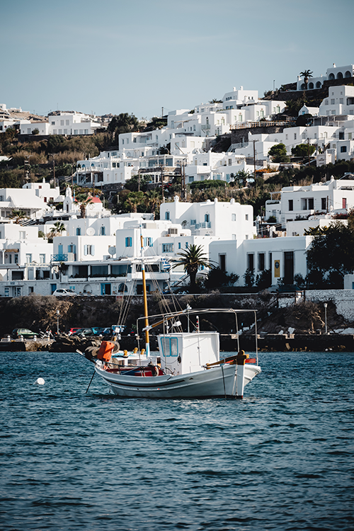 Mikonos'un ikonik bembeyaz evlerinin yeşilliklerle bezeli bir tepede sıralandığı büyüleyici Ege manzarasında, berrak masmavi sularda sallanan geleneksel bir balıkçı teknesi, bu seyahat rehberi için adanın özgün atmosferini yansıtıyor.