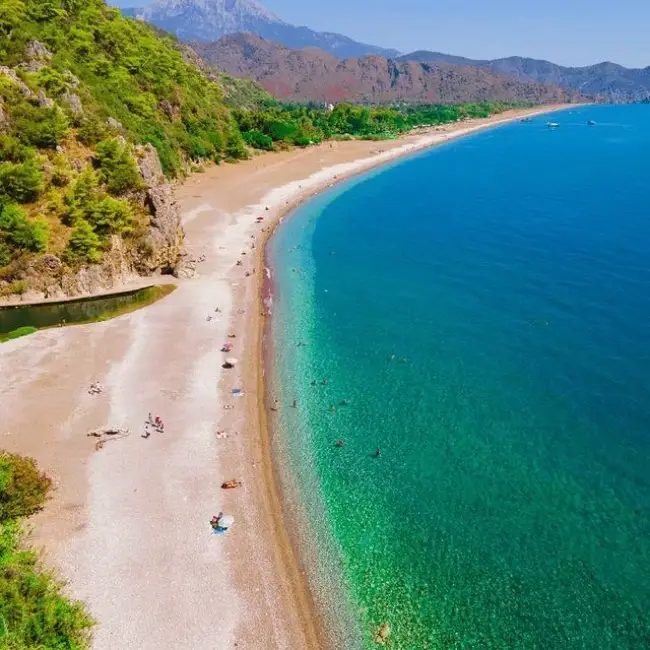 Uzaktan çekilmiş havadan bir görüntüde, yemyeşil tepelerle çevrili, kristal berraklığında turkuaz sulara sahip uzun ve kıvrımlı Olimpos Plajı, denizde yüzen ve kumsalda güneşlenen insanlarla birlikte Antalya'nın en güzel plajlarından biri olarak görünüyor.