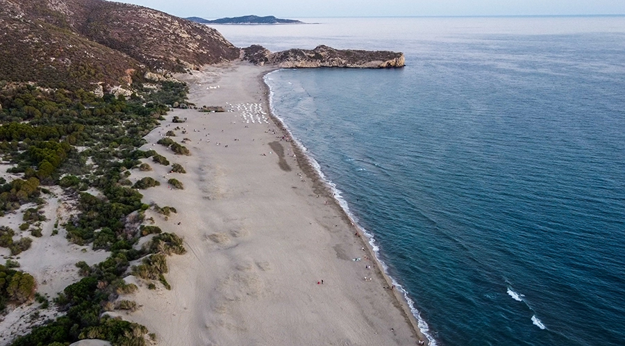 Patara Plajı'nın kuşbakışı görünümünde, geniş ve uzun kumlu sahil boyunca masmavi Akdeniz suları hafif dalgalarla kıyıya vururken, sağda kayalık bir burun denize uzanıyor ve kum tepeleri ile çevrili yeşil bitki örtüsü sol tarafta uzanarak Antalya'nın en güzel plajlarından birinin doğal güzelliğini sergiliyor.