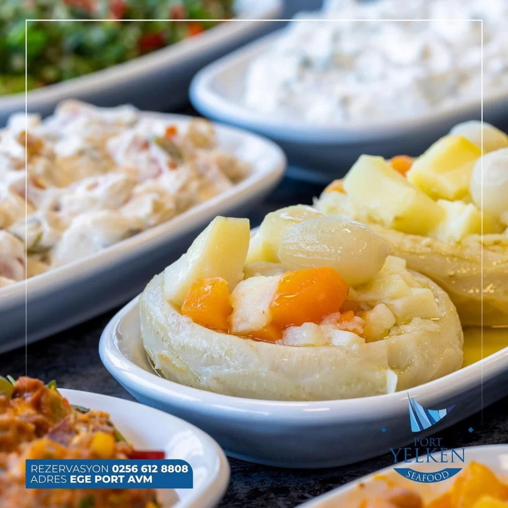 Ortada parlak zeytinyağı ile doldurulmuş, patates, arpacık soğan ve havuç parçalarıyla bezeli bir zeytinyağlı enginar tabağı, bulanık arka planda çeşitli meze çeşitleri ve ön planda Port Yelken Seafood logosuyla iştah açıcı bir sunum.