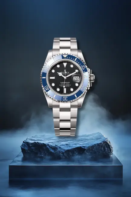 Mavi seramik bezeli ve parlak siyah kadranıyla öne çıkan, metal bilezikli bir Rolex Submariner dalış saati, denizin derinliklerinden ilham alan lacivert saat modellerini vurgulayan dumanlı koyu mavi bir ortamda, taş bir kaide üzerinde sunuluyor.