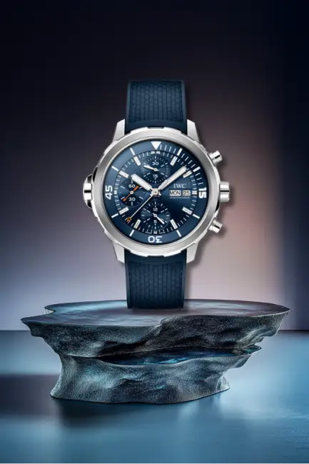 Derin lacivert seramik bezeli ve kadranıyla denizin mistik atmosferini yansıtan IWC Aquatimer Chronograph, paslanmaz çelik kasası, kronograf sayaçları ve tırtıklı kauçuk kayışıyla şık bir şekilde kaya benzeri bir platform üzerinde sergilenen, dalış tutkunları için ideal bir saat modelidir.