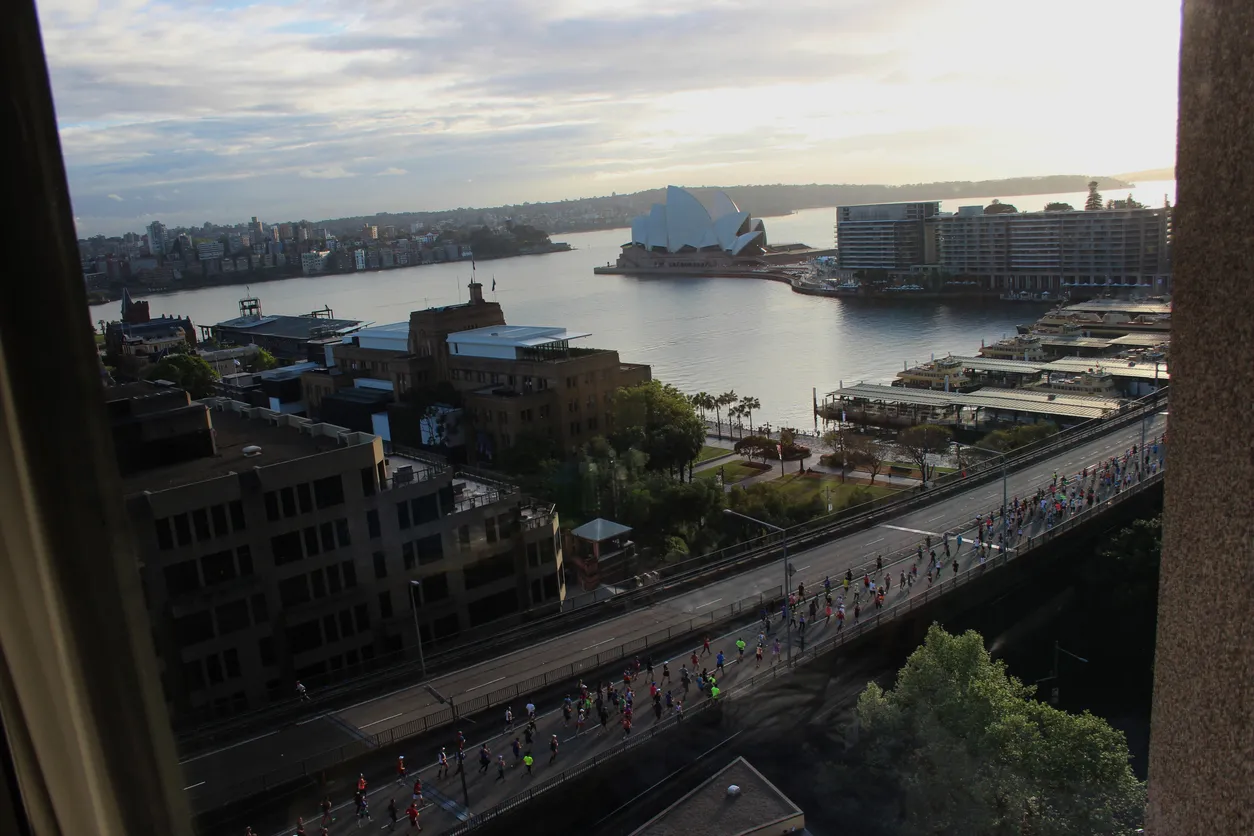 Güneşli bir Sidney sabahında, binlerce enerjik koşucunun ikonik Sidney Opera Binası ve Harbour Köprüsü'nün eşsiz silüeti önünden geçerek tarihi Sidney Maratonu parkurunda ilerlediği, coşkulu kalabalıkların tezahüratlarıyla dolu canlı bir sahne.
