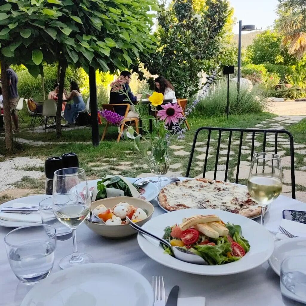 Bir bahçe restoranında, yeşillikler içindeki bir masada bol peynirli pizza, taze salatalar ve kadehlerde beyaz şarapların servis edildiği, arka planda ağaçların altında oturan insanların göründüğü keyifli bir açık hava yemeği anı.