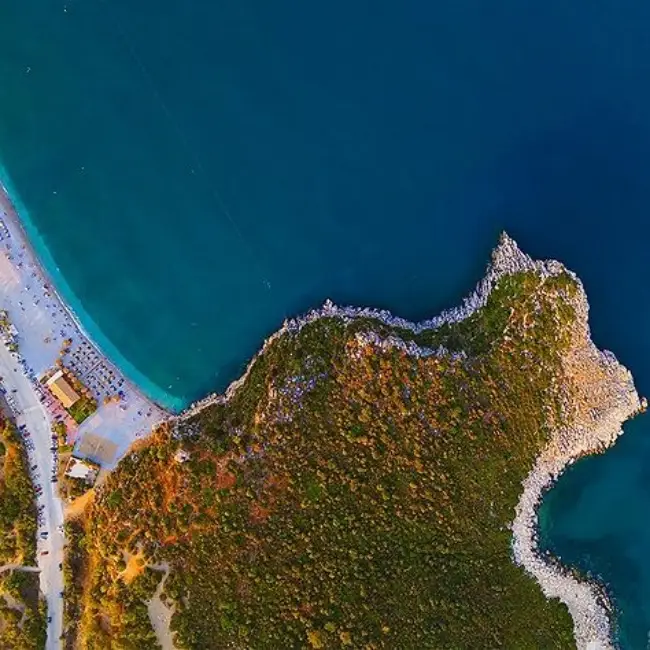 Demre Taşdibi Plajı'nın kuşbakışı görüntüsü, turkuaz sulara sahip geniş kumsalı, şemsiyeler ve şezlonglarla dolu sahil şeridi, yanındaki yemyeşil, kayalık burun ve masmavi Akdeniz'in huzurlu birleşimiyle Antalya'nın en güzel plajlarından biri olarak öne çıkıyor.