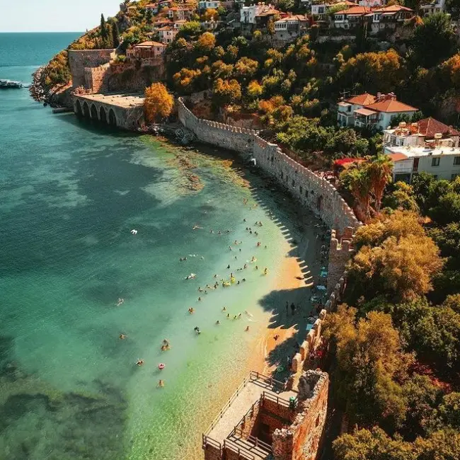 Tarihi Alanya Tersanesi'nin etkileyici taş duvarları boyunca uzanan, berrak turkuaz ve zümrüt yeşili sularında yüzen çok sayıda tatilciyle dolu, altın rengi ince kuma sahip Tersane Plajı, Antalya'nın en güzel plajlarından biri olarak parlıyor ve çevresindeki yeşil yamaçlardaki yerleşim yerleriyle doğal ve tarihi güzelliği bir araya getiriyor.