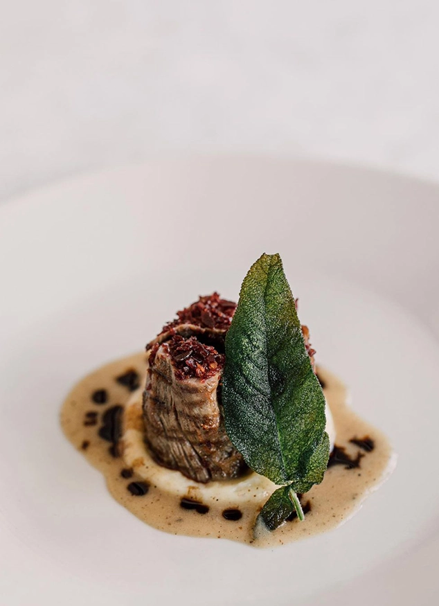 Urla'nın Bağ Rotaları boyunca keşfedilebilecek zengin gastronomisini yansıtan, beyaz bir tabağın ortasında, etrafı benekli kahverengi sosla çevrili püre üzerinde servis edilmiş, üstü kızıl renkli bir harçla zenginleştirilmiş et yemeği ve taze adaçayı yaprağıyla süslenmiş zarif bir sunum.