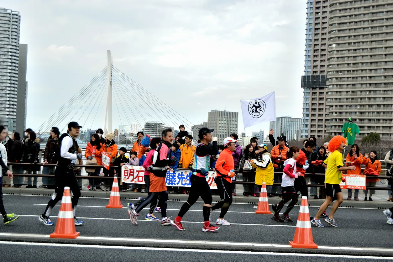 Tokyo Maratonu'nda yarışan binlerce koşucunun, şehrin canlı atmosferinde ve tarihi binalarının önünden geçerken yollar boyunca dizilen coşkulu seyircilerin desteğiyle azimle ilerlediği, spor ve dayanıklılık ruhunu yansıtan dinamik bir an.